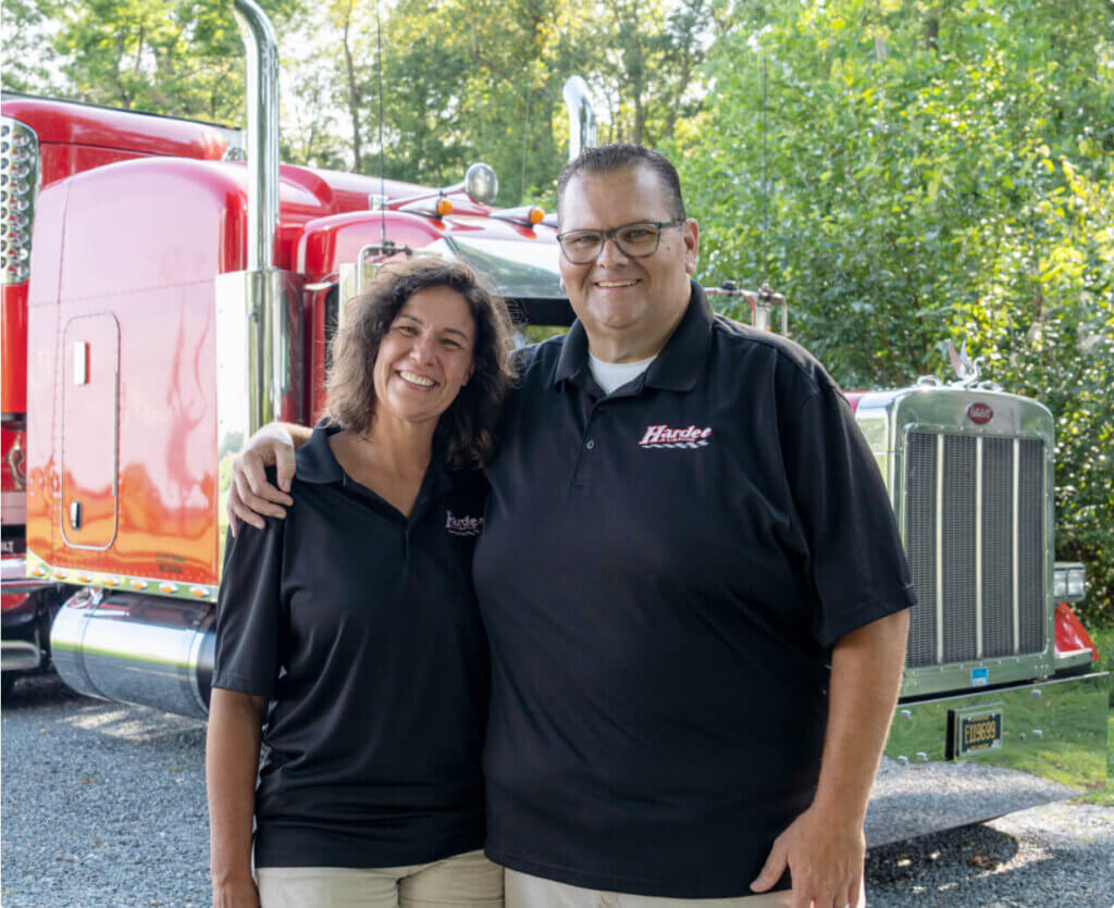 owner-operator-lori-and-wayne-hardee-standing-together-in-front-of-reefer-freight-hauler-truck-looking-at-camera